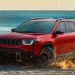 Red Jeep Cherokee SUV driving across sandy terrain near the beach, kicking up sand while showcasing its bold front grille, black wheels, roof rails, and adventurous capability with the ocean in the background.