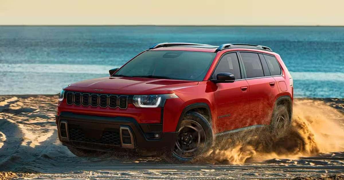 Red Jeep Cherokee SUV driving across sandy terrain near the beach, kicking up sand while showcasing its bold front grille, black wheels, roof rails, and adventurous capability with the ocean in the background.
