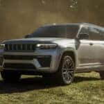 Silver Jeep Grand Cherokee SUV parked on a grassy field in natural sunlight, highlighting its refined exterior design, signature seven-slot grille, premium wheels, and spacious SUV profile with trees in the background.