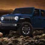 Blue Jeep Wrangler Rubicon pickup truck parked on a rocky riverbed at sunset, showcasing its rugged off-road design, large all-terrain tires, and signature seven-slot grille against a mountainous landscape.