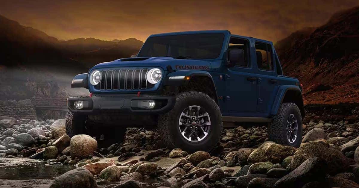 Blue Jeep Wrangler Rubicon pickup truck parked on a rocky riverbed at sunset, showcasing its rugged off-road design, large all-terrain tires, and signature seven-slot grille against a mountainous landscape.