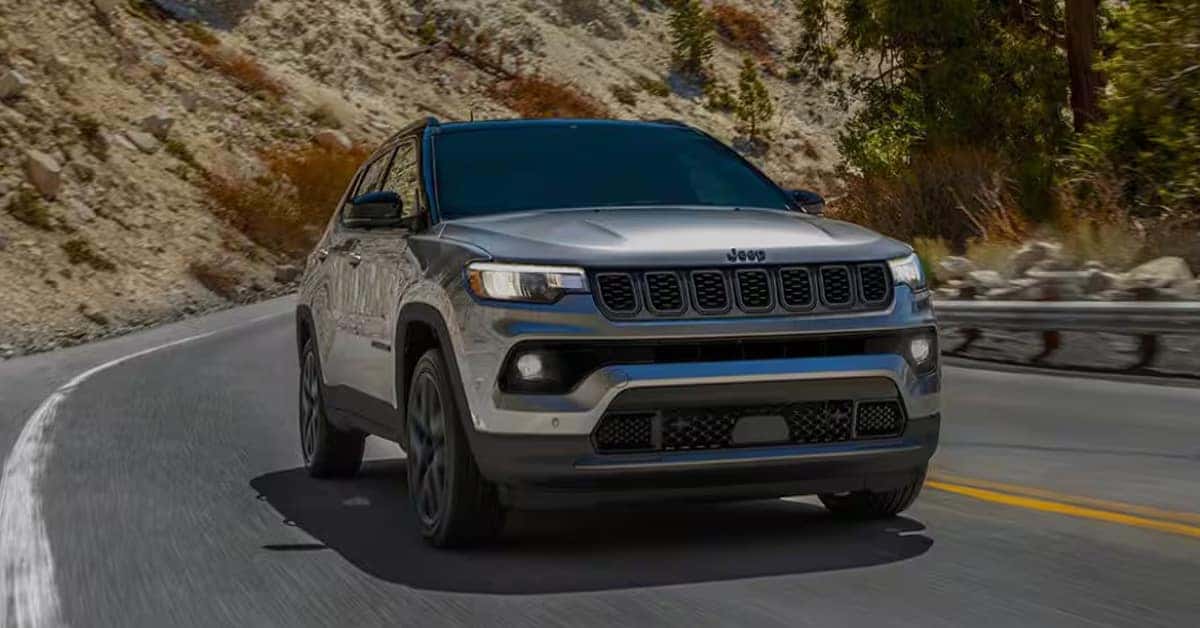 Silver Jeep Compass driving on a winding mountain road with LED headlights illuminated and rocky hillsides in the background.