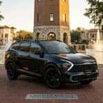 A professional, golden hour photograph of a black 2026 Kia Sportage HEV featuring the new vertical Star Map amber LED signature, parked on a brick plaza at Cranes Roost Park in Altamonte Springs, Florida, with the illuminated town tower and fountains visible in the background.