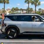 A silver Kia EV9 plugged into a red Tesla Supercharger station with a sign noting cross-compatibility for 2026+ models in a sunny shopping center parking lot.