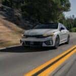 Low-angle front view of a 2026 Kia K5 navigating a curve on a scenic road.