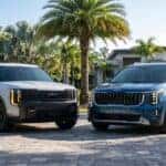 A white 2027 Kia Telluride X-Pro and a blue 2026 Kia Carnival parked side-by-side in a luxurious, sunlit Florida driveway lined with palm trees.