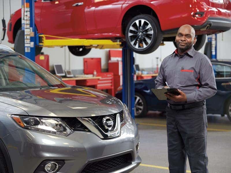 Nissan Service Technician A mechanic standing next to a silver Nissan with a clipboard