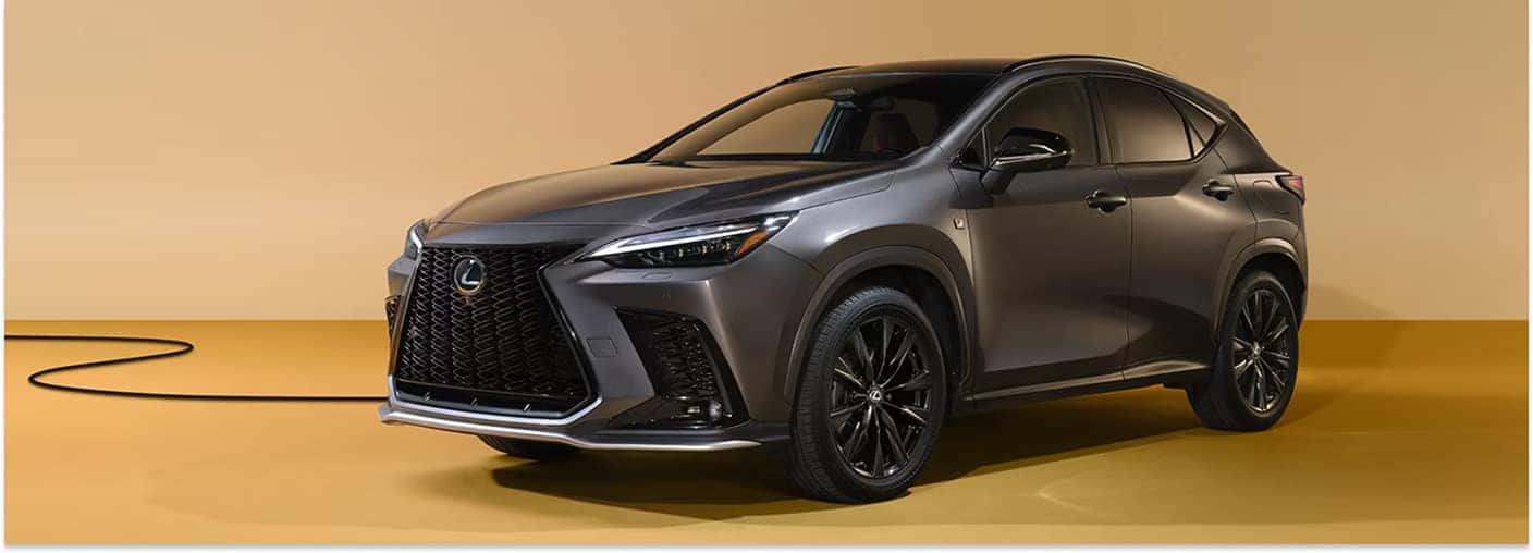 > Front three-quarter view of a dark metallic gray 2026 Lexus NX Hybrid with a prominent spindle grille and black multi-spoke wheels, positioned in a studio setting against a solid tan background with a black cable on the floor.