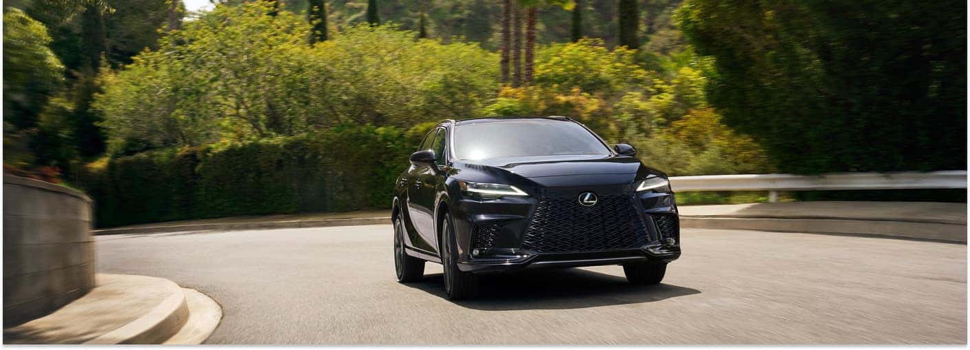 > Front three-quarter view of a black 2026 Lexus RX driving through a curve on an asphalt road bordered by lush green trees and foliage under bright sunlight.