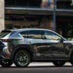 A blue-gray Mazda CX-5 drives forward against a city building.