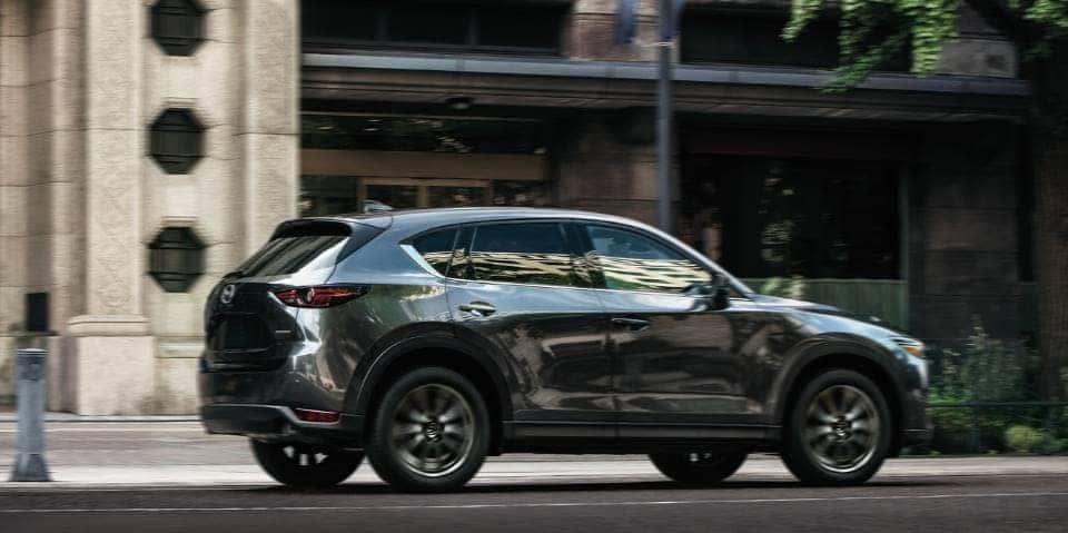 A blue-gray Mazda CX-5 drives forward against a city building.