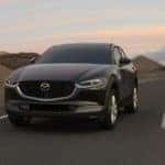 A gray 2021 Mazda CX-30 driving towards the camera on an empty road. Small mountains and a sunset in the background.