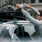 a brunette man in a gray shirt with a leather watch and a silver wedding ring and sunglasses loading luggage into a cargo net roof rack on a mazda SUV.
