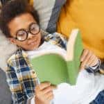 little boy wearing glasses and laying on the couch while reading a book