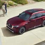 A 2025 Mazda CX-90 PHEV parked in front of a modern building, surrounded by lush greenery and rocky terrain.