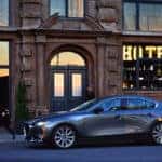A Mazda 3 sedan parked in front of a modern hotel, showcasing its sleek design and vibrant color against the backdrop.