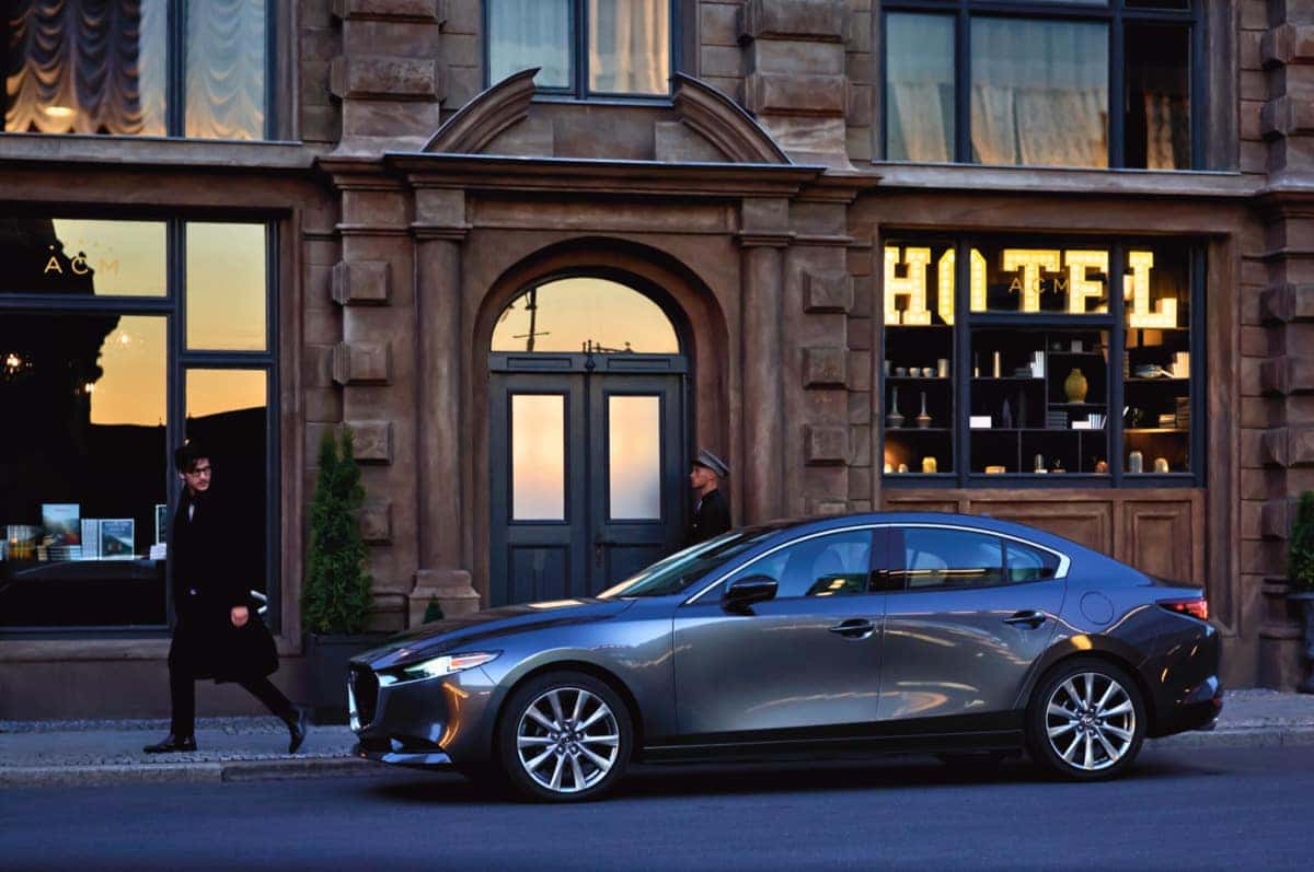A Mazda 3 sedan parked in front of a modern hotel, showcasing its sleek design and vibrant color against the backdrop.