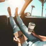 Woman holding coffee and a map, feet propped on dashboard on a summer road trip
